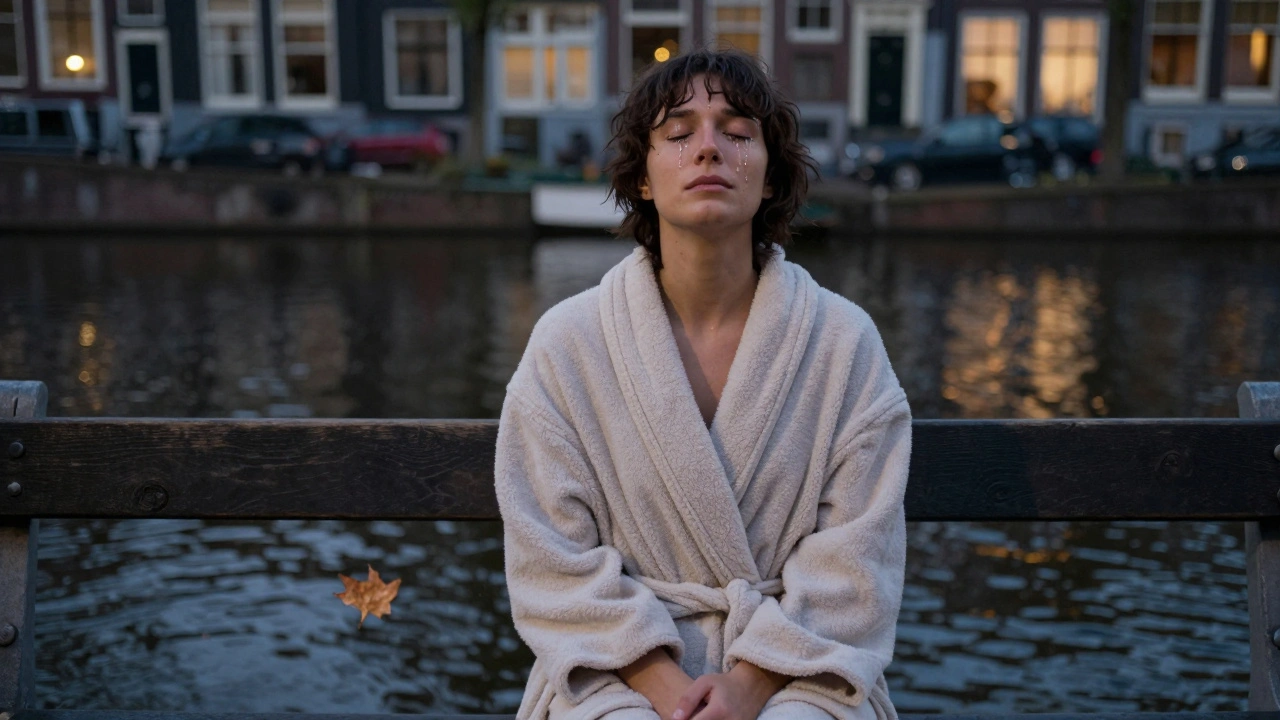 A person sitting alone by a canal at dusk, wrapped in a robe, crying softly in quiet emotional release.